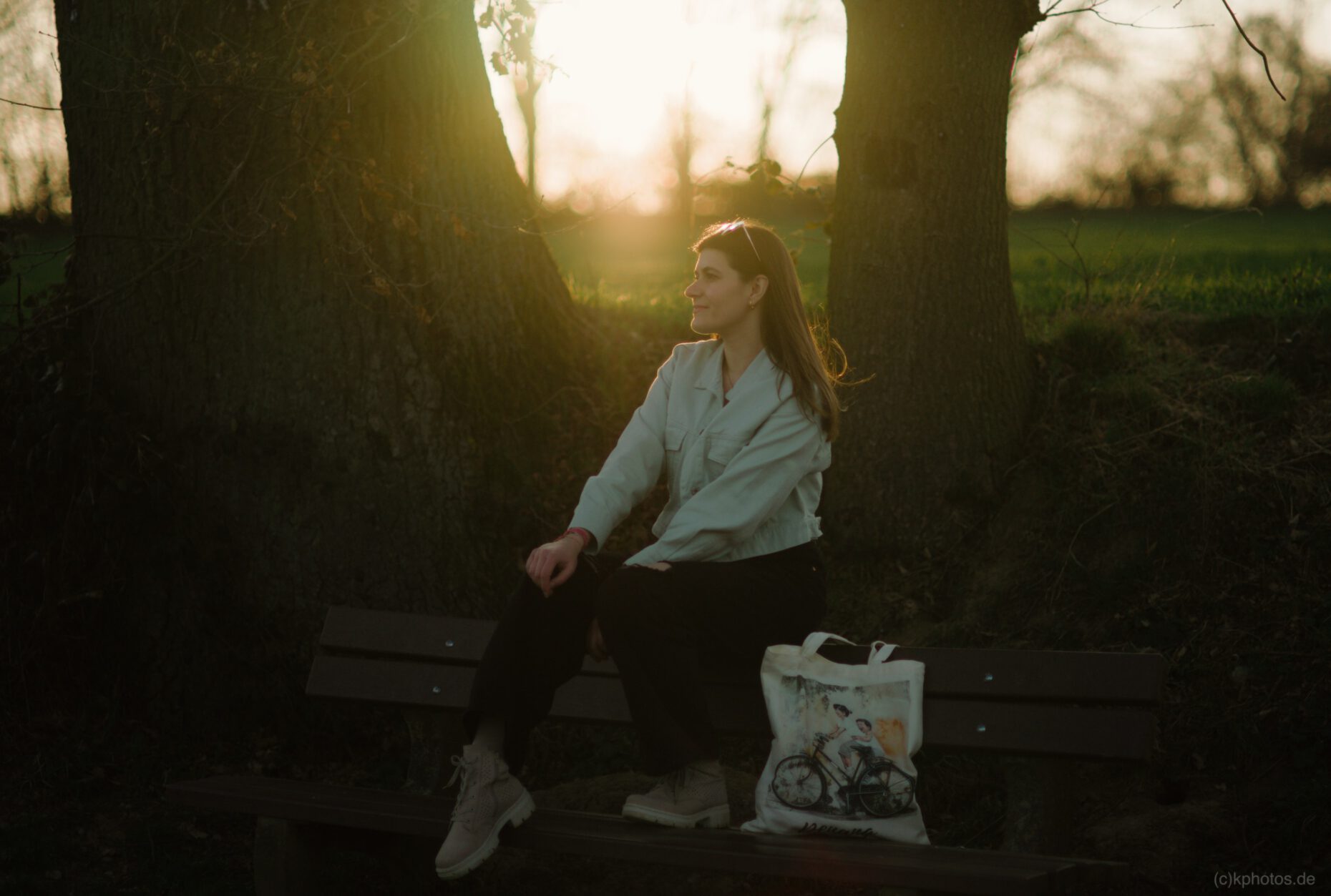 A woman is sitting on a park bench, with trees in the background and the sun setting in the distance